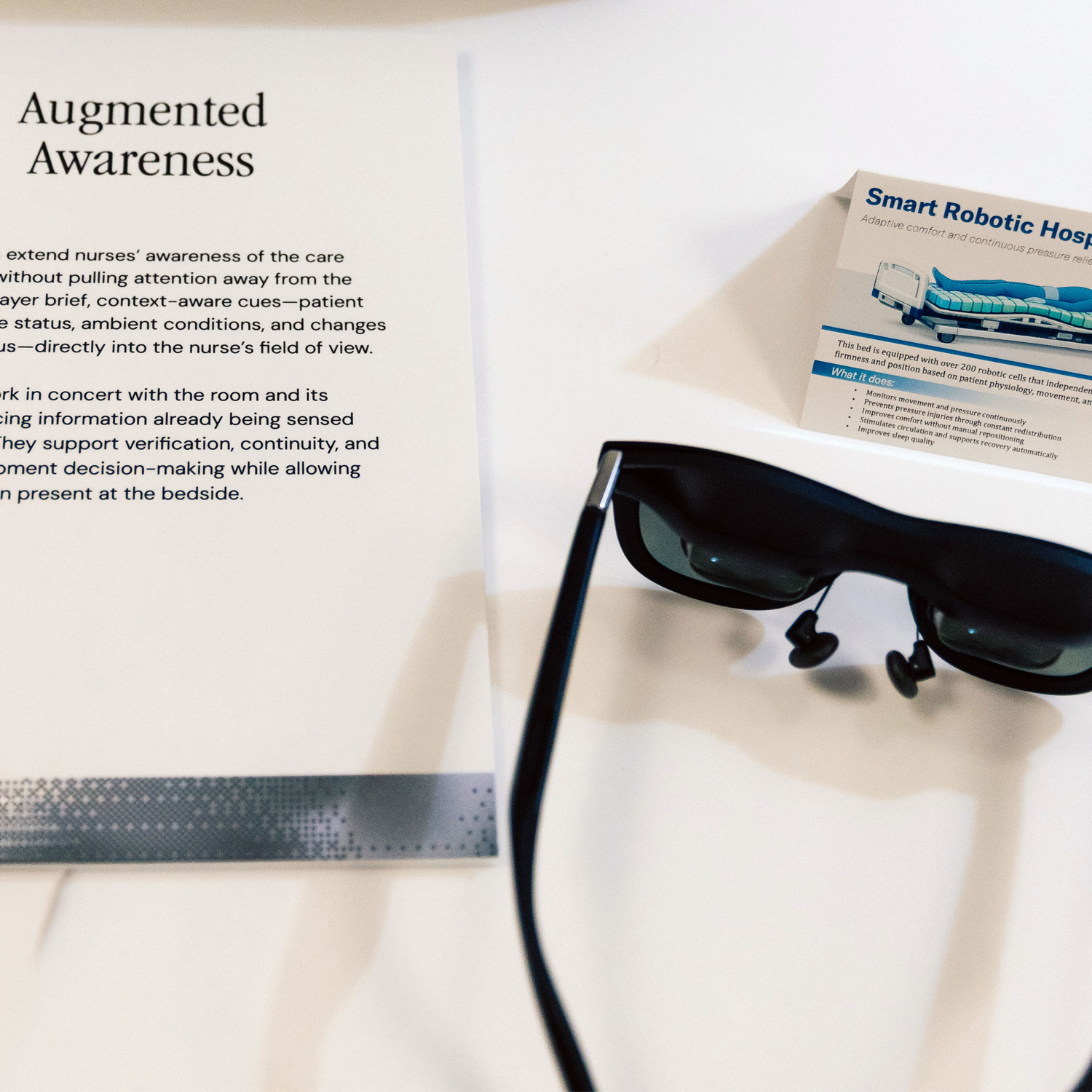 An augmented-reality eyepiece resting on a placard reading &lsquo;Augmented Awareness,&rsquo; next to a Smart Robotic Hospital Bed information card &mdash; a quiet artifact moment from inside the prototype.