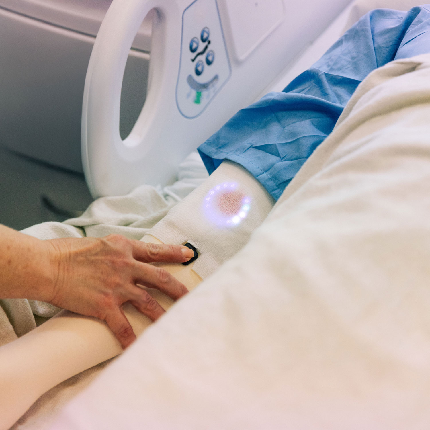 A nurse&rsquo;s hand rests on a patient&rsquo;s arm; an illuminated smart bandage glows softly above the wound site, monitoring the dressing without interrupting human touch.